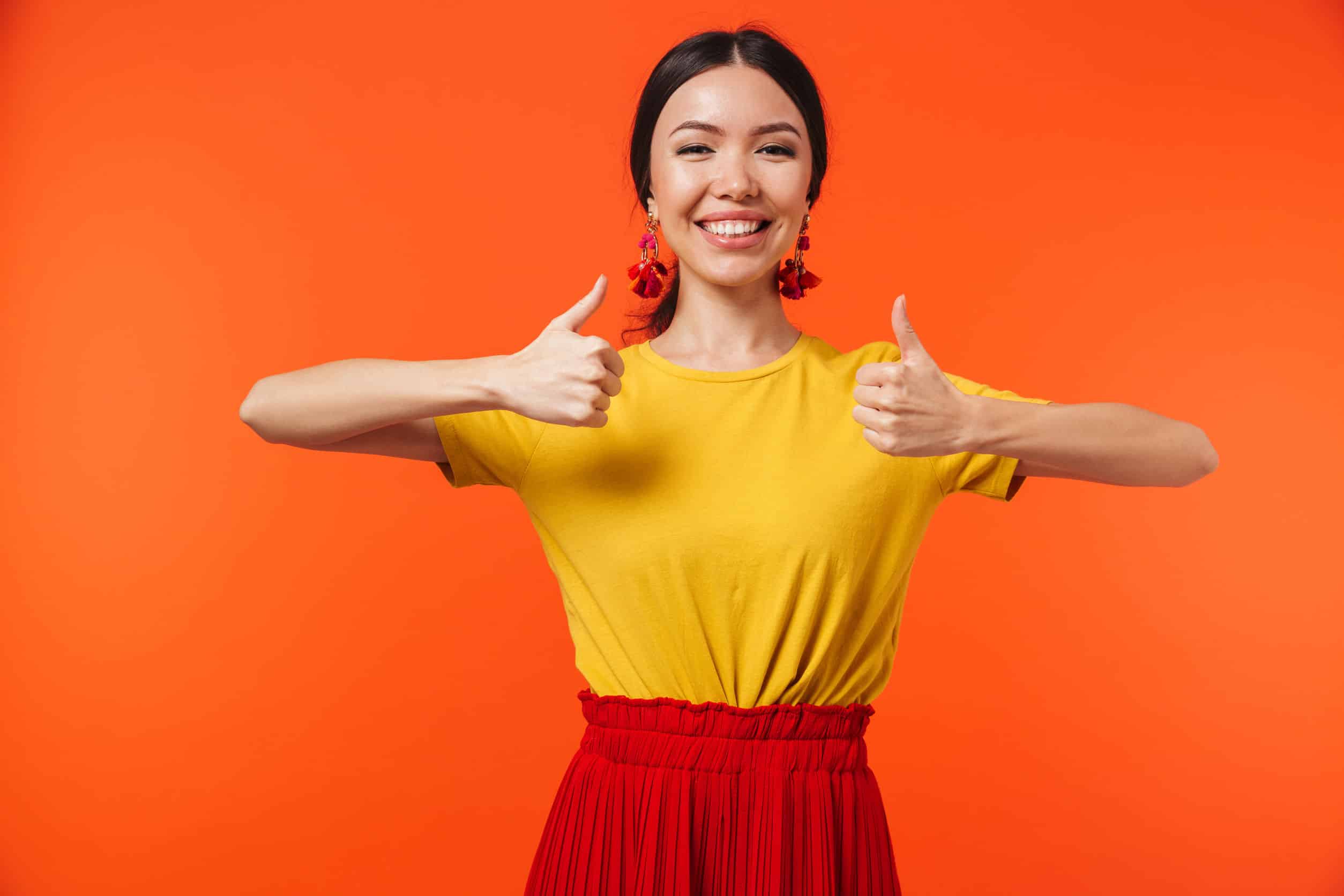 a happy young woman with two thumbs up on against an orange wall background