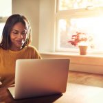 Woman smiling while drinking coffee and sitting with her laptop in the kitchen