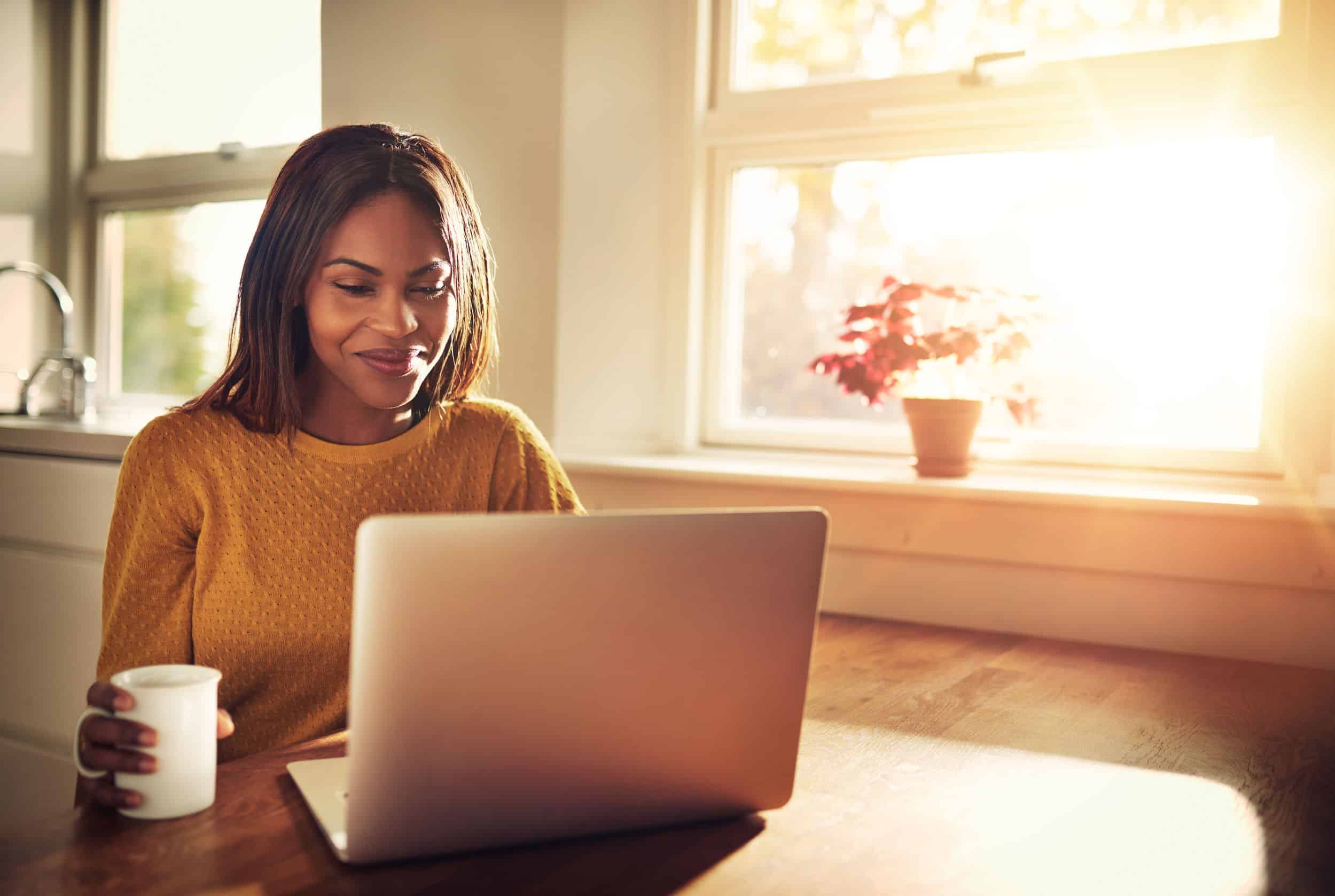 Woman smiling while drinking coffee and sitting with her laptop in the kitchen