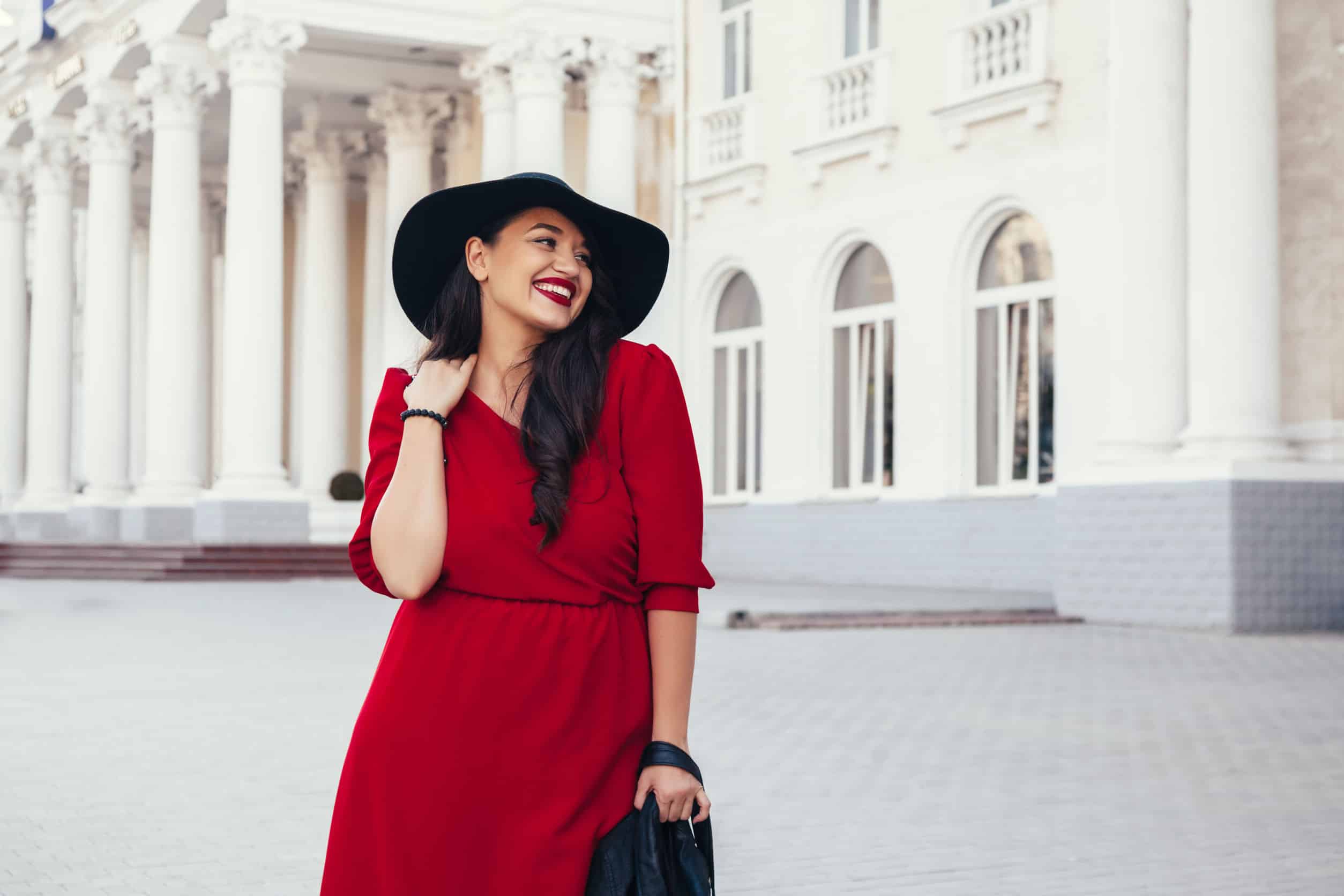 smiling brunette woman with red dress and lipstick outside a mansion