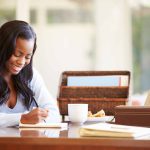A woman writing in a notebook at her desk with a cup of coffee, a snack, a plant, and a basket of files
