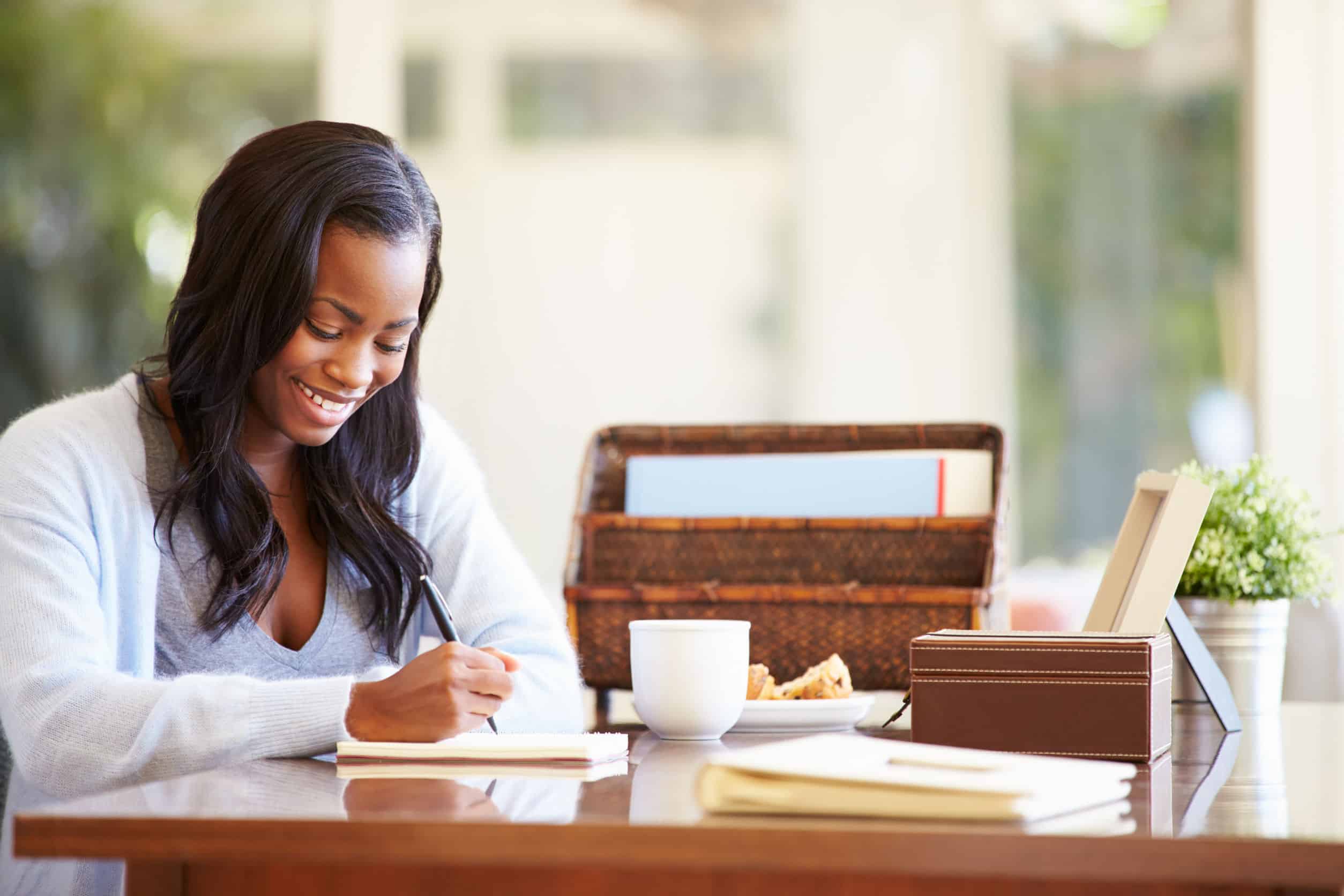 A woman writing in a notebook at her desk with a cup of coffee, a snack, a plant, and a basket of files