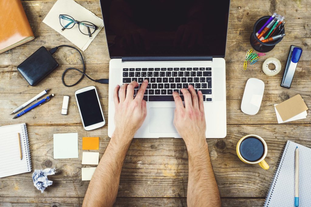 A man writing on a laptop with notebooks, coffee, pens, and post-it notes on his desk